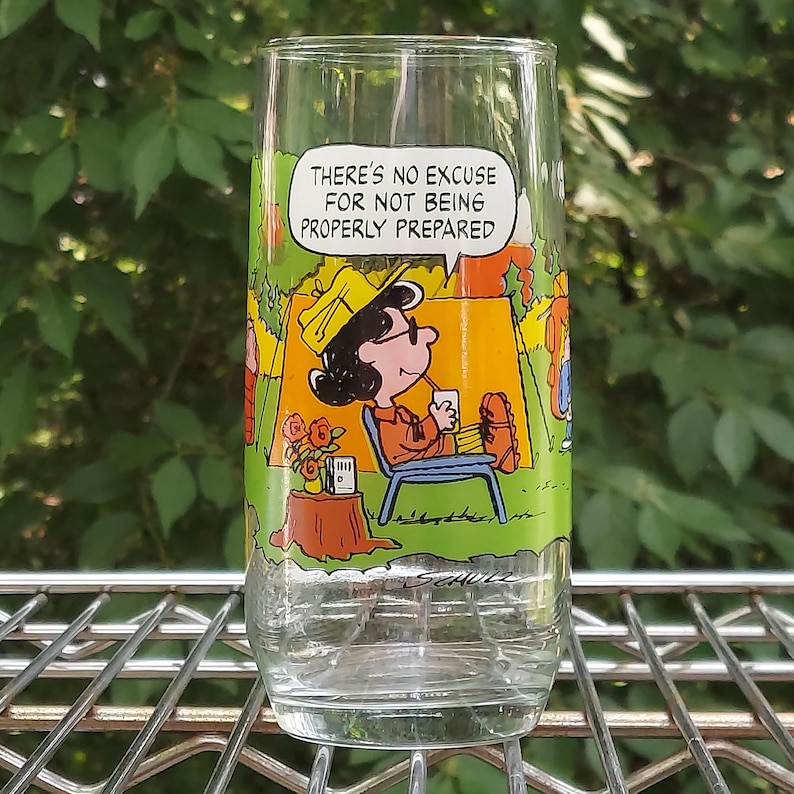 May include: A clear glass featuring a cartoon illustration of a character from the Peanuts comic strip. The character is seated in a camping chair, with a speech bubble that reads, "THERE'S NO EXCUSE FOR NOT BEING PROPERLY PREPARED."