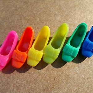 Puede incluir: Una colección de seis mini zapatos de tacón de plástico en colores del arcoíris: rosa, naranja, amarillo, verde y azul. Cada zapato tiene una correa decorativa en la parte superior. Los zapatos están dispuestos en fila.
