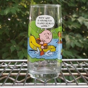 May include: A clear glass tumbler featuring a cartoon illustration of Charlie Brown in a yellow boat, paddling on a river. The glass has the text "RATS! WHY IS HAVING FUN ALWAYS SO MUCH WORK?" and the artist's signature.
