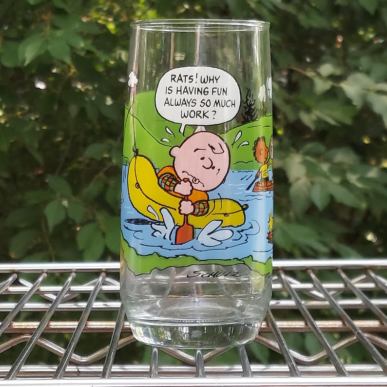 May include: A clear glass tumbler featuring a cartoon illustration of Charlie Brown in a yellow boat, paddling on a river. The glass has the text "RATS! WHY IS HAVING FUN ALWAYS SO MUCH WORK?" and the artist's signature.
