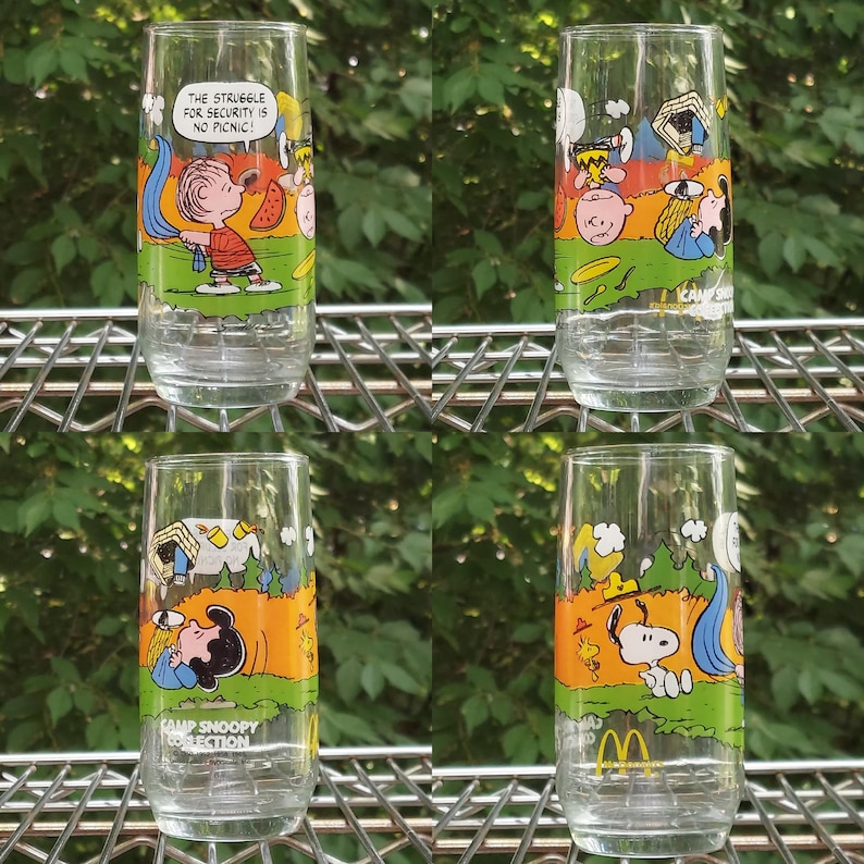 May include: Four clear glass tumblers with colorful cartoon illustrations of Peanuts characters. Each glass features a different scene with characters like Charlie Brown and Snoopy. One glass has the text "THE STRUGGLE FOR SECURITY IS NO PICNIC!"