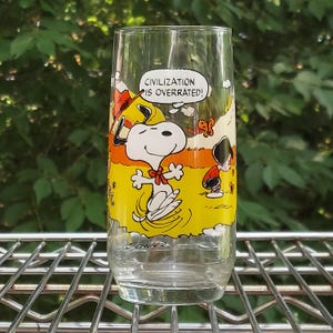 May include: A clear glass featuring a cartoon image of Snoopy with the text "CIVILIZATION IS OVERRATED!". The glass has a yellow and orange background with Snoopy and a small figure. The glass is sitting on a metal rack.
