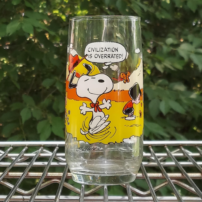 May include: A clear glass featuring a cartoon image of Snoopy with the text "CIVILIZATION IS OVERRATED!". The glass has a yellow and orange background with Snoopy and a small figure. The glass is sitting on a metal rack.