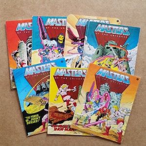 May include: A collection of vintage "Masters of the Universe" comic books. The covers feature vibrant illustrations of characters and scenes from the series, with bold text and dynamic layouts. The comics are in various colors, including red, blue, and yellow.