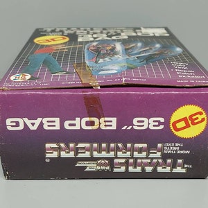 May include: A purple and yellow box for a 36 inch Transformers Bop Bag. The box features a 3D image of a Transformer character and the text "The Transformers More Than Meets The Eye!"  The box also includes the text "Heavy Duty Vinyl Repair Patch Included" and "Made in Taiwan".