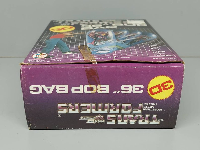 May include: A purple and yellow box for a 36 inch Transformers Bop Bag. The box features a 3D image of a Transformer character and the text "The Transformers More Than Meets The Eye!"  The box also includes the text "Heavy Duty Vinyl Repair Patch Included" and "Made in Taiwan".