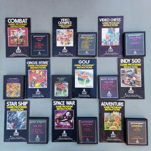 Op de afbeelding: Een verzameling Atari-gamecartridges en instructiehandleidingen, met titels als "Combat", "Video Olympics" en "Adventure". De covers tonen kleurrijke graphics en het Atari-logo, met klassieke videogamekunst.