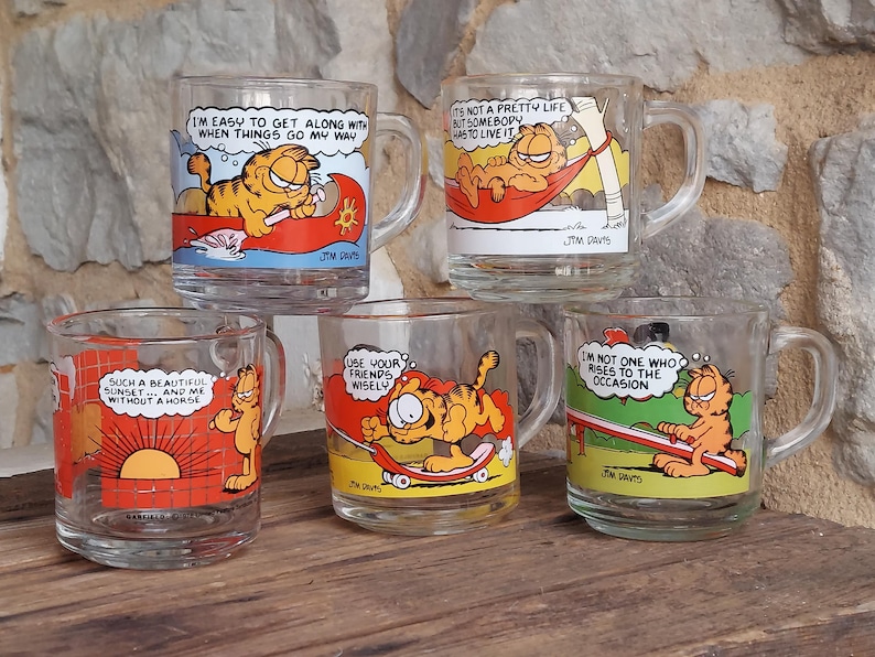May include: Set of five glass mugs featuring Garfield the cat in various humorous situations. Each mug has a different quote from Garfield, including "I'm easy to get along with when things go my way", "It's not a pretty life but somebody has to live it", "Such a beautiful sunset...and me without a horde", "Use your friends wisely", and "I'm not one who rises to the occasion".