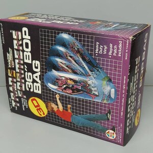 May include: A Transformers 36-inch inflatable bop bag with a 3D image of a robot. The bag is purple, blue, and black. The box is black with white text and a picture of a child kicking the bag.