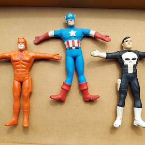 May include: Three superhero action figures: Daredevil in orange, Captain America in blue, red, and white, and The Punisher in black and white. The figures are posed with arms outstretched, showcasing their iconic costumes and designs.