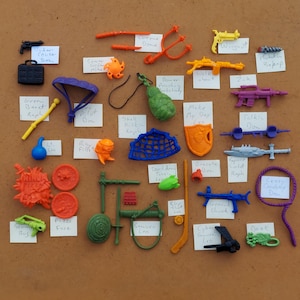 Puede incluir: Una colección de juguetes de plástico de colores, que incluyen un gorro verde, un paracaídas morado, una red azul, un escudo rojo, una pistola amarilla, una tortuga verde, una pizza roja y naranja y una espada samurái verde. Los juguetes están dispuestos sobre una superficie marrón con etiquetas escritas a mano que identifican cada artículo.