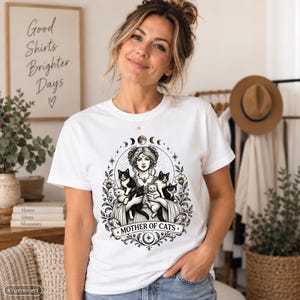 May include: A white short-sleeved t-shirt featuring a black and white graphic print of a person surrounded by five cats. The design includes floral elements, celestial symbols, and the text "MOTHER OF CATS" centered at the bottom of the illustration.