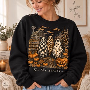 Könnte beinhalten: Ein schwarzes Sweatshirt mit Rundhalsausschnitt und einer Grafik von drei Geistern auf einem Feld mit Scheune, Kürbissen und Maispflanzen. Der Text lautet PUMPKINS, HAY RIDES, CORN MAZE und tis the season. Die Geister haben Karomuster, Leopardenmuster und Punkte.