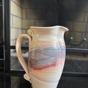 May include: A ceramic pitcher with a cream-colored base and a curved handle. The body features a horizontal band of blended blue, gray, and reddish-pink glaze across the center, contrasting with the smooth, light-toned surface of the vessel.