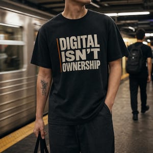 May include: A black short-sleeved t-shirt featuring the text "DIGITAL ISN'T OWNERSHIP" printed in large, light-colored capital letters on the chest, accompanied by a thin vertical orange line to the left of the first two lines of text.