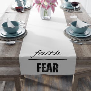 以下が含まれることがあります： 白いテーブルランナー。水平線の上に黒い筆記体で「faith」、その下に掠れた黒いブロック体で「FEAR」という文字がプリントされており、食器が並べられた木製のダイニングテーブルの中央に配置されている。
