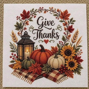 Può includere: Un ricamo a punto croce su tessuto bianco con il testo Give Thanks in caratteri neri. Il disegno include una lanterna, una grande zucca arancione, una zucca verde, una zucca bianca, girasoli, foglie autunnali e una coperta a quadri.