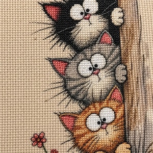 May include: A cross-stitch embroidery featuring three stacked cats peeking from behind a textured tree trunk. The top cat is black, the middle is gray, and the bottom is orange. Each cat has large white eyes, pink ears, and long whiskers.