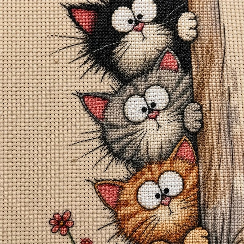 May include: A cross-stitch embroidery featuring three stacked cats peeking from behind a textured tree trunk. The top cat is black, the middle is gray, and the bottom is orange. Each cat has large white eyes, pink ears, and long whiskers.