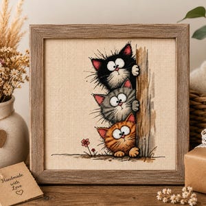 May include: A square wooden frame containing a cross-stitch embroidery on beige fabric. The design features three cartoon cats stacked vertically, peeking from behind a wooden post. The cats are black, gray, and orange with wide eyes.