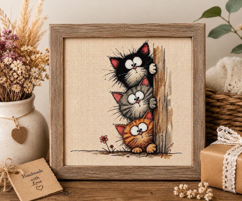 May include: A square wooden frame containing a cross-stitch embroidery on beige fabric. The design features three cartoon cats stacked vertically, peeking from behind a wooden post. The cats are black, gray, and orange with wide eyes.