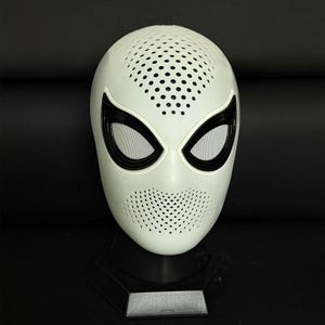 May include: A white full-face mask featuring a perforated texture on the forehead and chin areas. The mask has large, almond-shaped eye openings outlined in thick black frames with white mesh lenses, mounted on a black display stand.