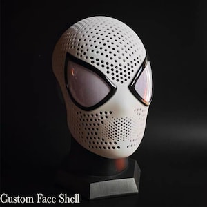 Peut inclure: Un masque de type coque faciale blanc présentant un motif dense de petites perforations circulaires sur toute la surface. Le masque comprend deux grandes ouvertures oculaires en amande bordées de noir avec des mailles fines sur les yeux.