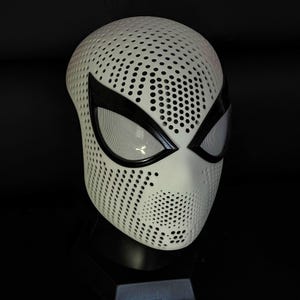 Può includere: Una maschera bianca che copre l'intera testa, caratterizzata da un fitto motivo di piccole perforazioni circolari su tutta la superficie. La maschera presenta due grandi aperture oculari a mandorla con bordi neri spessi e lenti a rete.