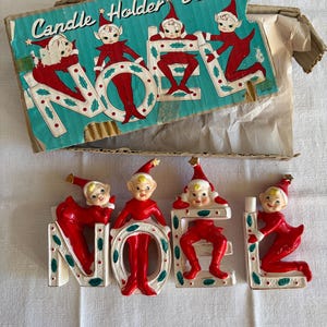 May include: A vintage ceramic candle holder set spelling NOEL, featuring four elf figurines in red outfits with gold accents. The set includes a cardboard box with the text Candle Holder Set and illustrations of the elves matching the figures.