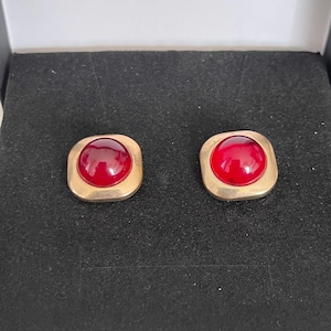May include: A pair of stud earrings featuring a smooth, rounded, deep red cabochon stone set within a square-shaped, brushed gold-tone metal frame. The earrings have a polished finish on the red center and a matte texture on the surrounding metal.