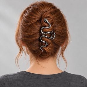 Puede incluir: Una pinza para el cabello de metal plateado con forma de serpiente enroscada. El accesorio presenta una superficie lisa y reflectante con una cabeza de serpiente detallada en la parte superior y un cuerpo sinuoso que sirve de cierre.