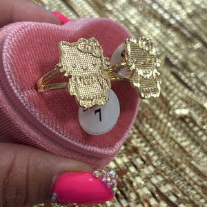 May include: Two gold-toned rings featuring a textured, flat-faced character design with a bow on the head. The rings are displayed on a pink velvet heart-shaped box, with one ring showing a small white circular tag printed with the number 7.
