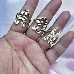 May include: Three gold-toned rings featuring cursive letters A, D, and M. Each ring has a polished, textured surface with diagonal etched lines across the metal. The rings are designed with an open, script-style font and a shiny, reflective finish.