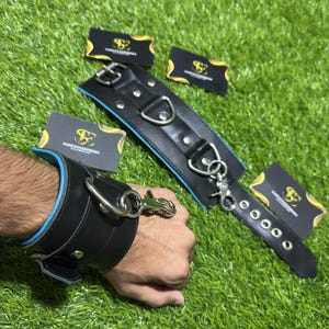 May include: A black leather cuff with a bright blue inner lining, featuring silver-toned metal hardware including a D-ring, a swivel snap hook, and a buckle closure with five adjustment holes. The cuff is accompanied by four small black cards.
