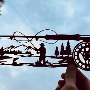 Puede incluir: Una pieza de arte mural de metal negro con la silueta de una escena de pesca con mosca. El diseño incluye una caña de pescar con carrete, una persona de pie en el agua lanzando el sedal, un pez saltando, pinos y picos de montañas al fondo.