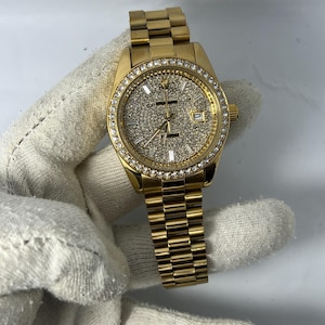 May include: A gold-tone wristwatch featuring a round case with a diamond-encrusted bezel and a matching gold-tone link bracelet. The dial is fully paved with small sparkling stones, gold-tone hour markers, a date window, and gold-tone hands.