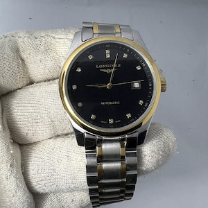 May include: A Longines automatic wristwatch featuring a black dial with diamond hour markers, a gold-tone bezel, and a two-tone silver and gold stainless steel link bracelet. The dial displays the Longines logo, the word AUTOMATIC, and a date window.