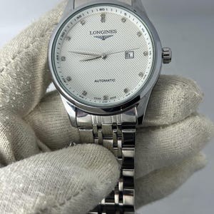 May include: A silver-tone Longines automatic wristwatch with a white textured dial, diamond hour markers, a date window at the three o'clock position, and a stainless steel link bracelet. The watch face features the Longines logo and text.