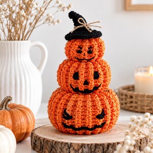 Peut inclure: Une décoration en forme de citrouilles au crochet empilées, composée de trois citrouilles orange de tailles différentes, chacune dotée de traits de visage noirs brodés. La citrouille du haut porte un petit chapeau de sorcière noir au crochet.