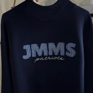 May include: A dark navy blue long-sleeve sweatshirt with a high mock neck collar. The front features the letters JMMS in a blue and white gingham check pattern, with the word patriots embroidered in white cursive script lettering below it.