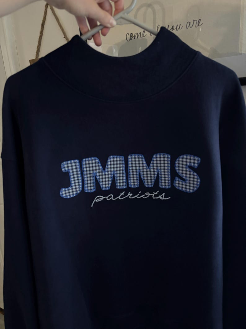 Op de afbeelding: Een donker marineblauw sweatshirt met lange mouwen en een hoge kraag. De voorzijde toont de letters JMMS in een blauw-wit vichy-ruitpatroon, met het woord patriots eronder geborduurd in witte cursieve letters.