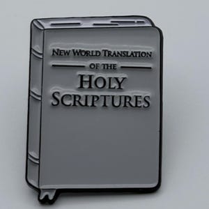 May include: A grey enamel pin shaped like a book with black outlines. The cover features the text "NEW WORLD TRANSLATION OF THE HOLY SCRIPTURES" in black lettering. The design includes a small bookmark tab at the bottom left corner of the book.
