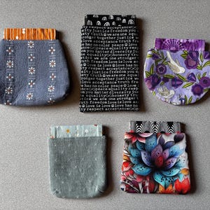 Peut inclure: Cinq pochettes en tissu aux motifs variés : une pochette grise à fleurs, une pochette noire avec du texte blanc incluant des mots comme truth et love, une pochette violette à fleurs, une pochette grise à croix blanches et une pochette à fleurs colorées.