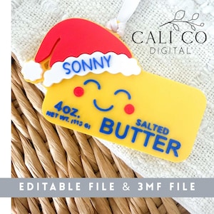 Könnte beinhalten: Ein gelbes Butterstück-Ornament mit einer rot-weißen Weihnachtsmütze. Das Gesicht zeigt einen lächelnden Ausdruck mit roten Wangen. Der Text lautet SONNY, 4OZ. NET WT. 113G und SALTED BUTTER. Das Design enthält eine weiße Schlaufe zum Aufhängen.