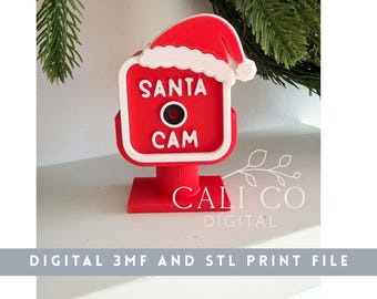 May include: A red and white 3D printed Santa Cam ornament featuring a square camera housing with a central black lens, topped with a Santa hat, and the text SANTA CAM in white lettering on the front, standing on a square base.