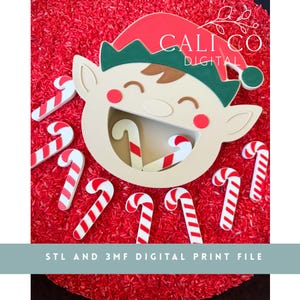 May include: A beige elf face cutout with a red and green hat, featuring a wide open mouth and red cheeks. Several small white and red striped candy cane shapes are scattered around the elf. Text reads CALI CO DIGITAL and STL AND 3MF DIGITAL PRINT FILE.
