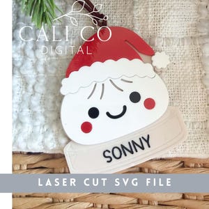 Puede incluir: Un adorno con forma de bola de masa blanca que lleva un gorro de Papá Noel rojo con borde blanco, ojos negros, una sonrisa negra y mejillas rojas. La base es una placa rectangular blanca con el nombre SONNY impreso en letras negras.