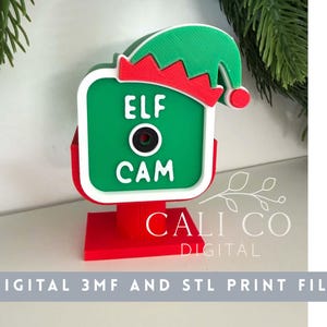 Puede incluir: Un letrero decorativo impreso en 3D con forma de cámara, con cara verde, borde blanco y base roja. Presenta un centro de lente circular negro y texto blanco que dice ELF CAM. Un gorro de elfo verde y rojo se sitúa en la esquina superior derecha.