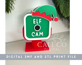 May include: A 3D printed decorative sign shaped like a camera with a green face, white border, and red base. It features a black circular lens center and white text reading ELF CAM. A green and red elf hat sits on the top right corner of the frame.