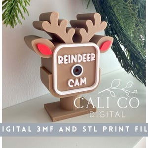 May include: A tan, 3D-printed reindeer-shaped camera housing featuring large antlers, red inner ears, and a central square frame with the white text REINDEER CAM surrounding a circular lens opening, mounted on a square base.